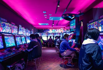 A vibrant arcade with rows of gaming machines adorned with colorful screens. Several people are immersed in playing games, seated on stools in a dimly lit room accented by an intense pink ceiling light. The atmosphere is lively and engaging.