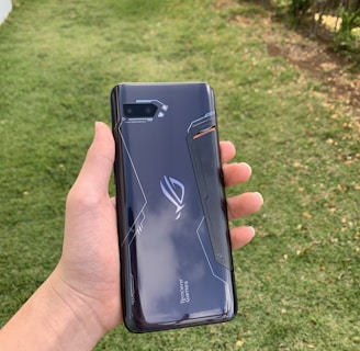 A hand holding a sleek, dark-colored smartphone with geometric patterns and the text 'Tencent Games' on the back cover. The background features a grassy area with some trees or bushes on the side.