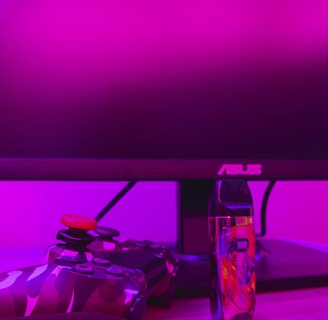 A gaming setup is featured with a camo-patterned game controller and a colorful vaporizer placed on a wooden desk. The background is illuminated with vibrant pink lighting, and a computer monitor with an ASUS logo is visible.