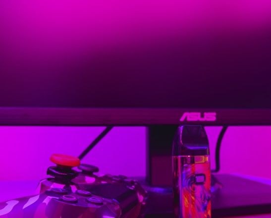 A gaming setup is featured with a camo-patterned game controller and a colorful vaporizer placed on a wooden desk. The background is illuminated with vibrant pink lighting, and a computer monitor with an ASUS logo is visible.