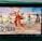 A handheld gaming console displays a screenshot of a fighting game featuring cartoonish characters in dynamic poses. The characters are standing in a virtual arena setting with a sky and cloud backdrop. The console is placed on a green surface.