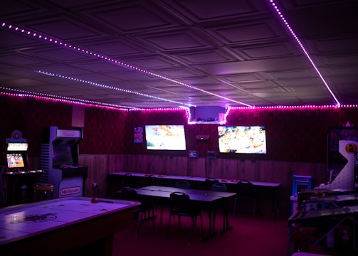 A dimly lit room with vibrant purple LED strip lights on the ceiling, featuring multiple gaming stations. There are two large screens showing video games, along with various arcade machines, including a Nintendo arcade cabinet. An air hockey table is positioned in the foreground.