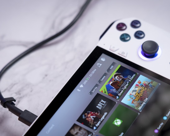 A handheld gaming console with a white body and colorful buttons features a screen displaying various game icons. The device is connected to a power source via a black cable and rests on a marble surface. The interface shows games like Apex and Street Fighter.