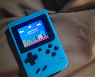 A handheld gaming device with a bright blue casing rests on a soft, dark fabric background. The screen displays a pixelated video game scene with options to select either Chinese or English language. The layout includes a classic D-pad and buttons, reminiscent of retro gaming consoles.