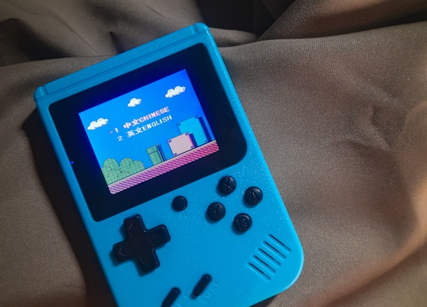 A handheld gaming device with a bright blue casing rests on a soft, dark fabric background. The screen displays a pixelated video game scene with options to select either Chinese or English language. The layout includes a classic D-pad and buttons, reminiscent of retro gaming consoles.